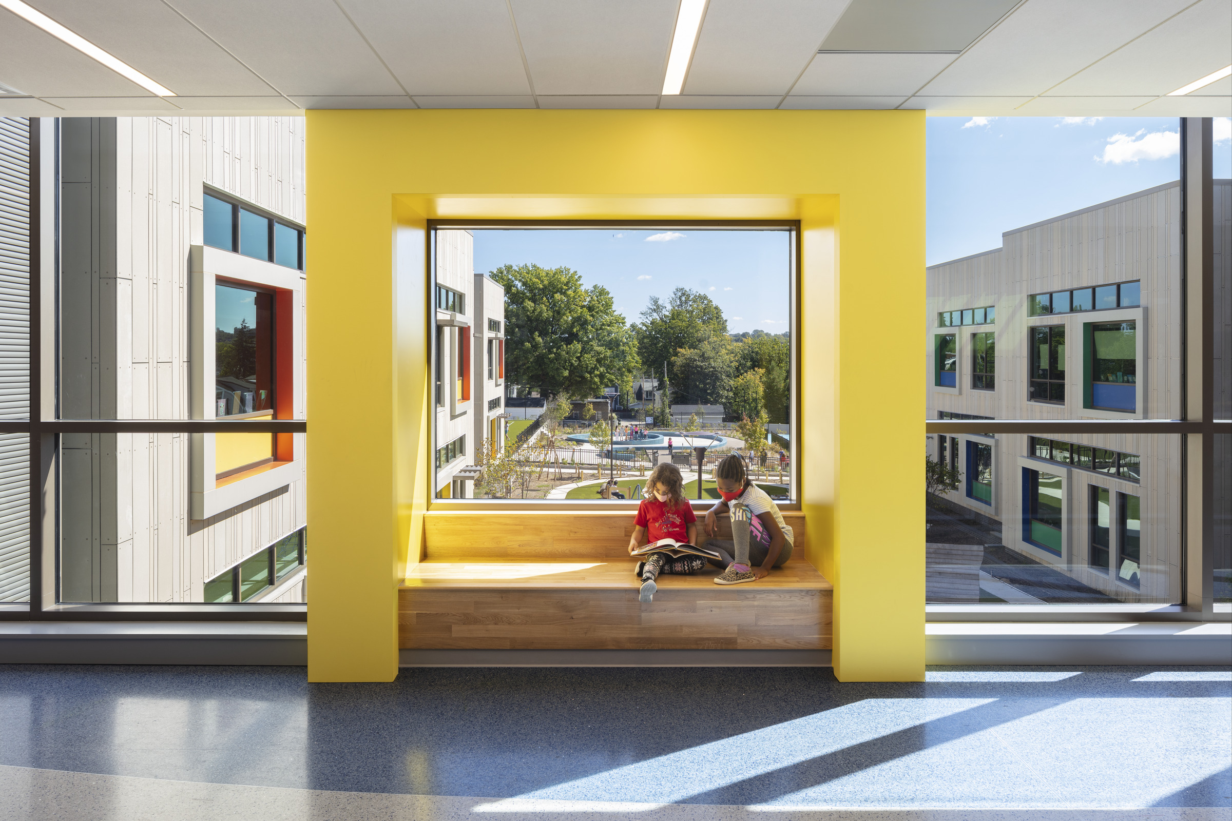 Children read a book on a bench in a brightly lit, sunny hallway of John Lewis Elementary School.