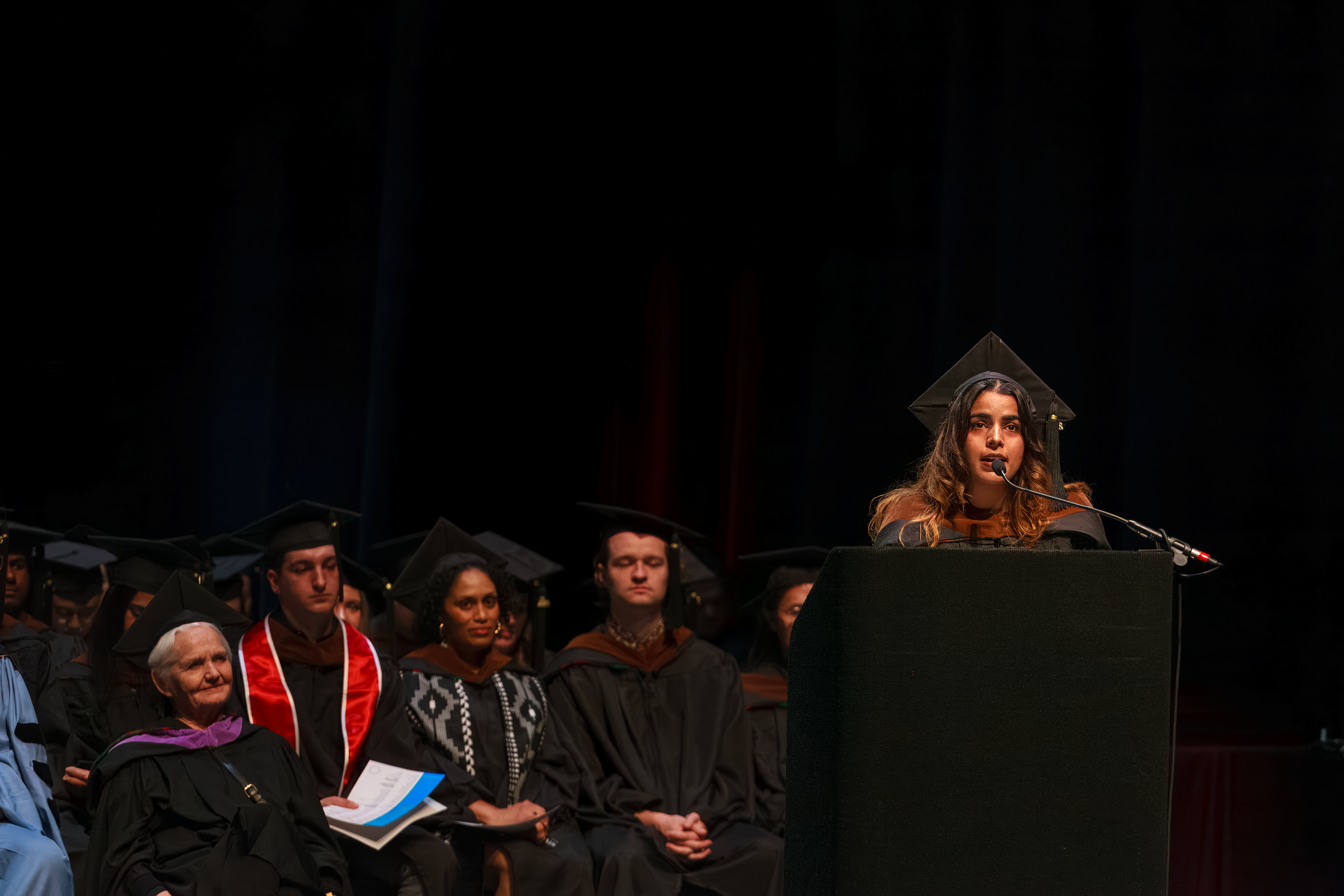 student at podium onstage in graduation regalia with others sitting in background
