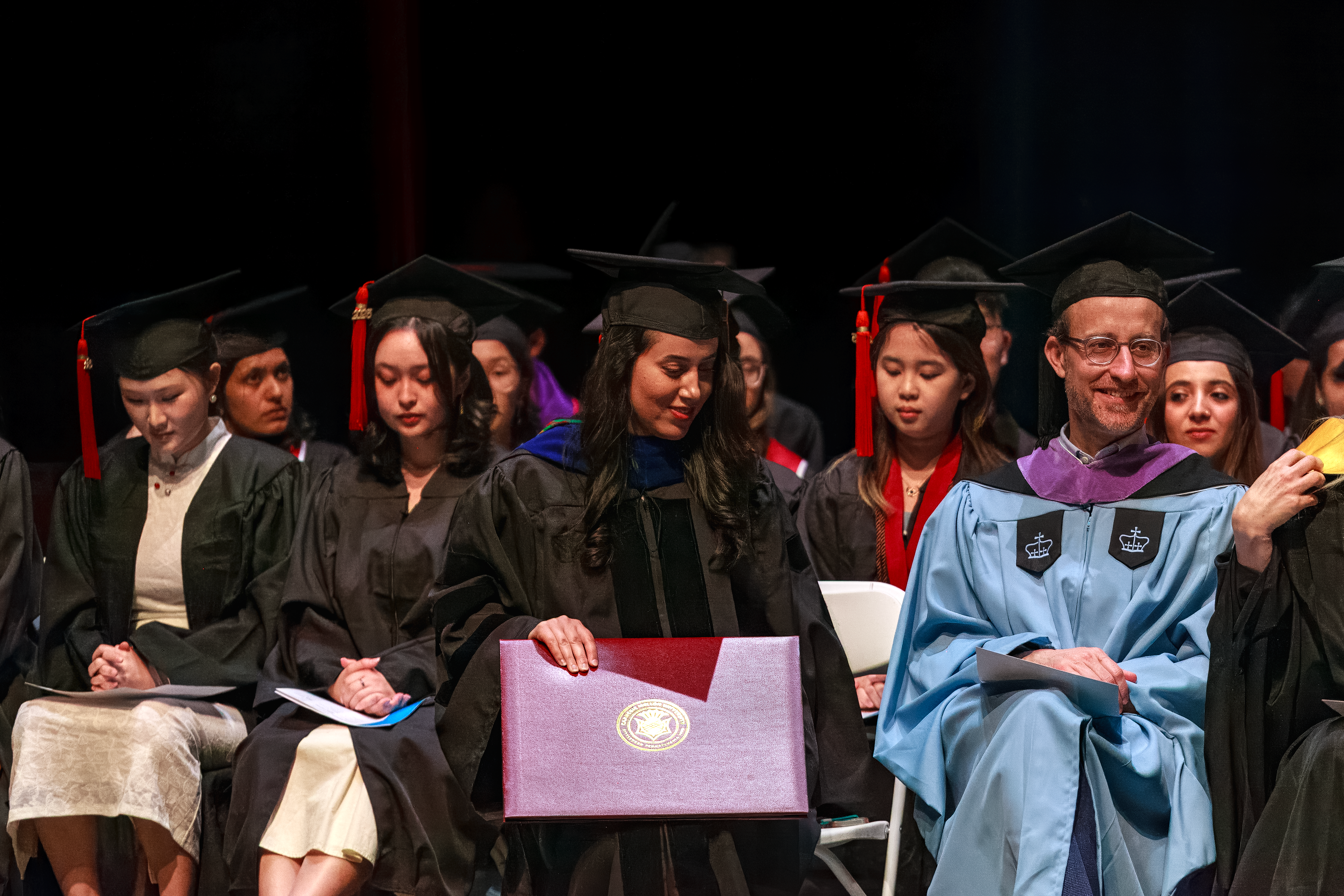 students and faculty sit onstage in graduation regalia.