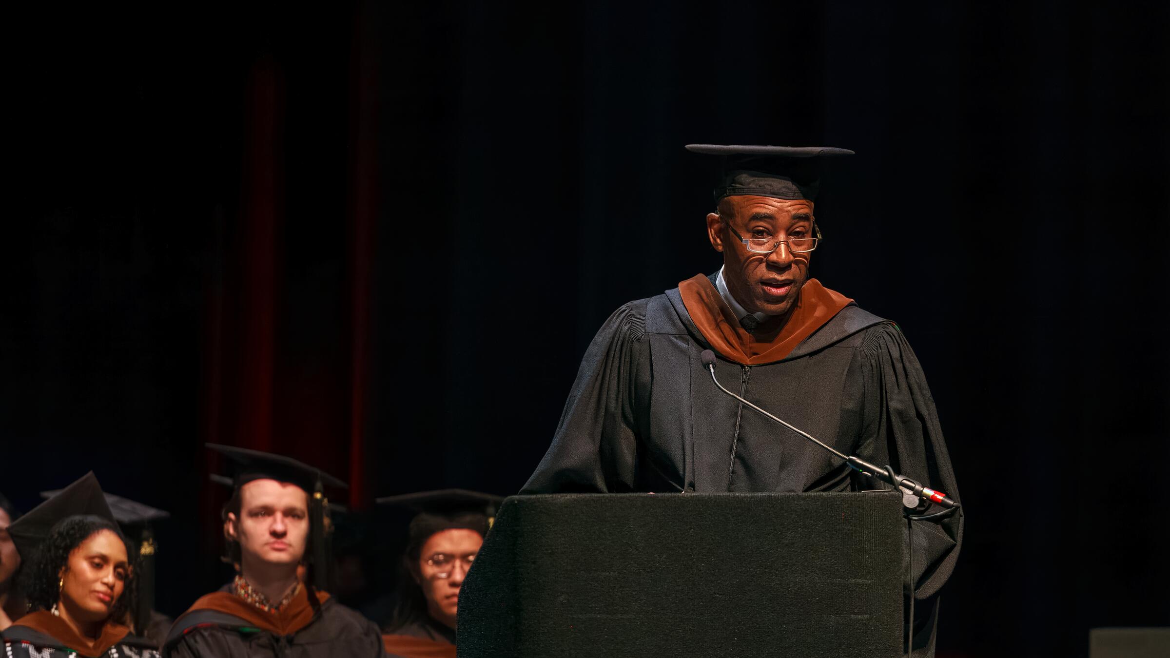 man speaking onstage in graduation regalia