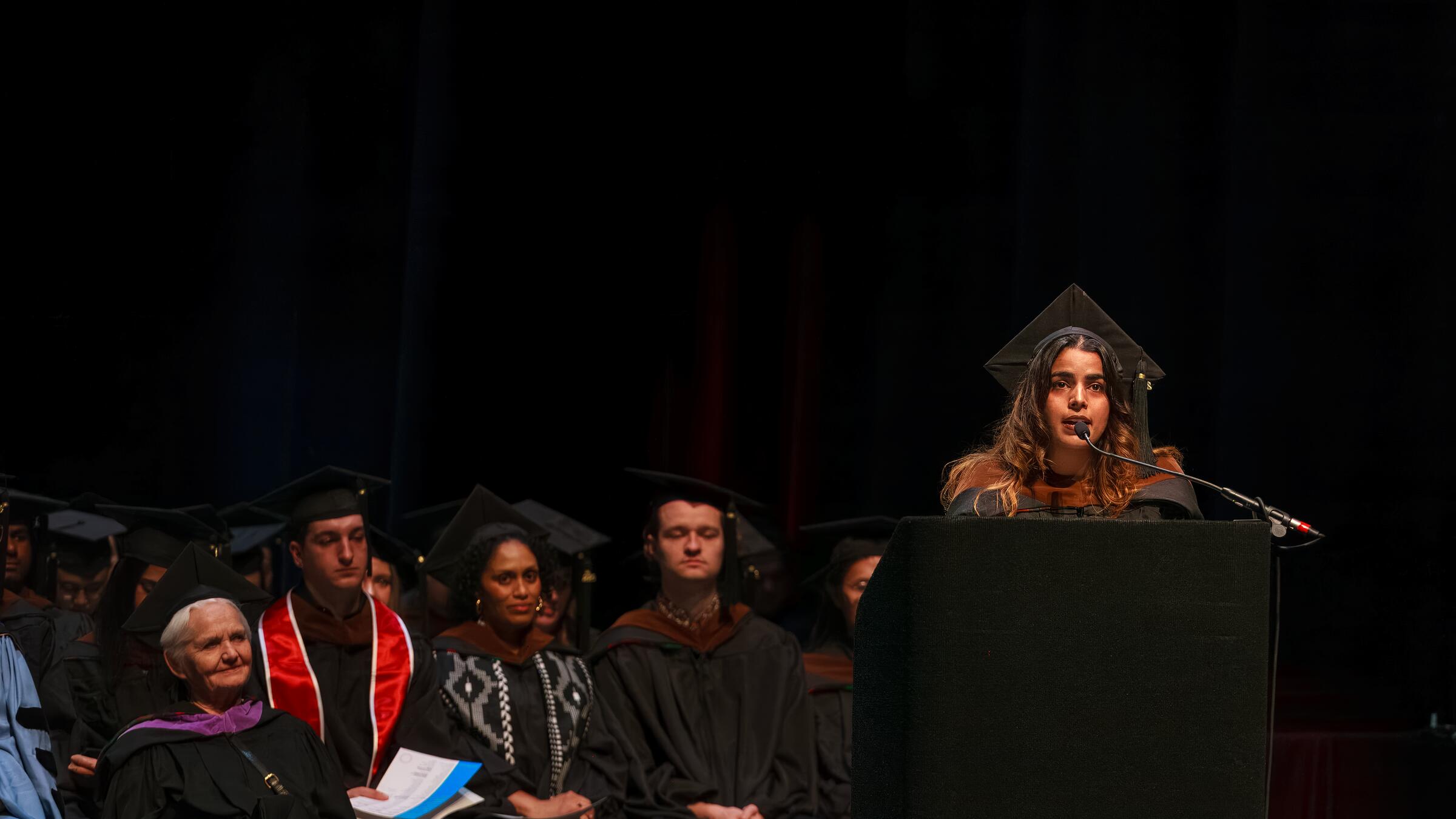 student at podium onstage in graduation regalia with others sitting in background