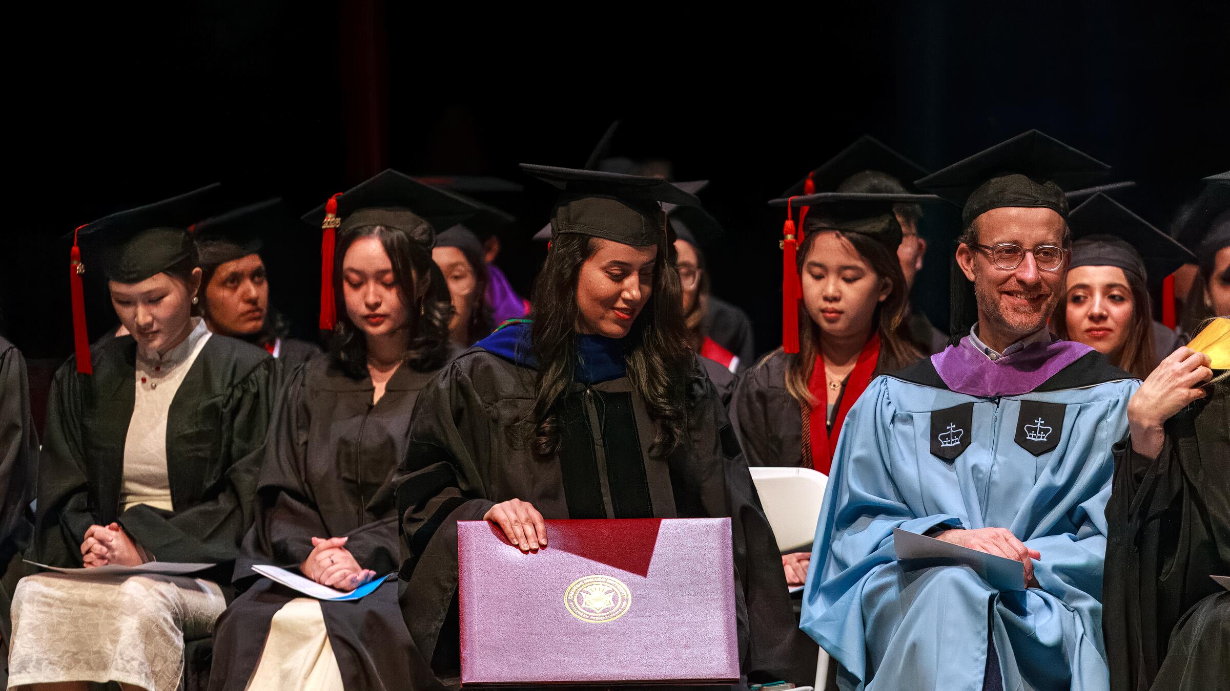 students and faculty sit onstage in graduation regalia.