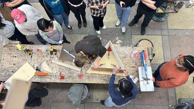 a group of students gathers around while two people build a triangular structure out of wood