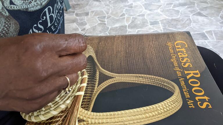 a hand weaves a sweetgrass basket on top of a book cover