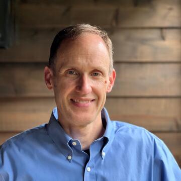 person with short hair wearing a blue collared shirt in front of a wooden slat wall