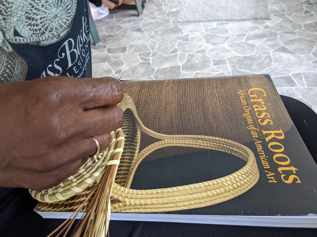 a hand weaves a sweetgrass basket on top of a book cover