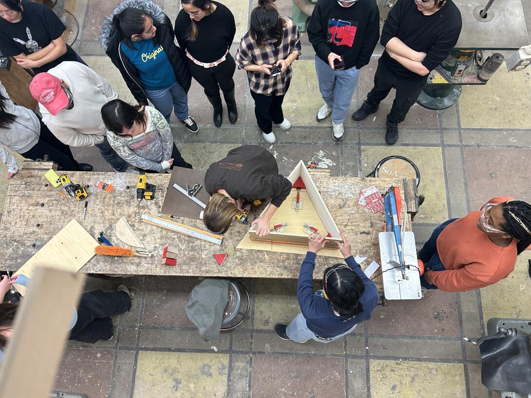 a group of students gathers around while two people build a triangular structure out of wood