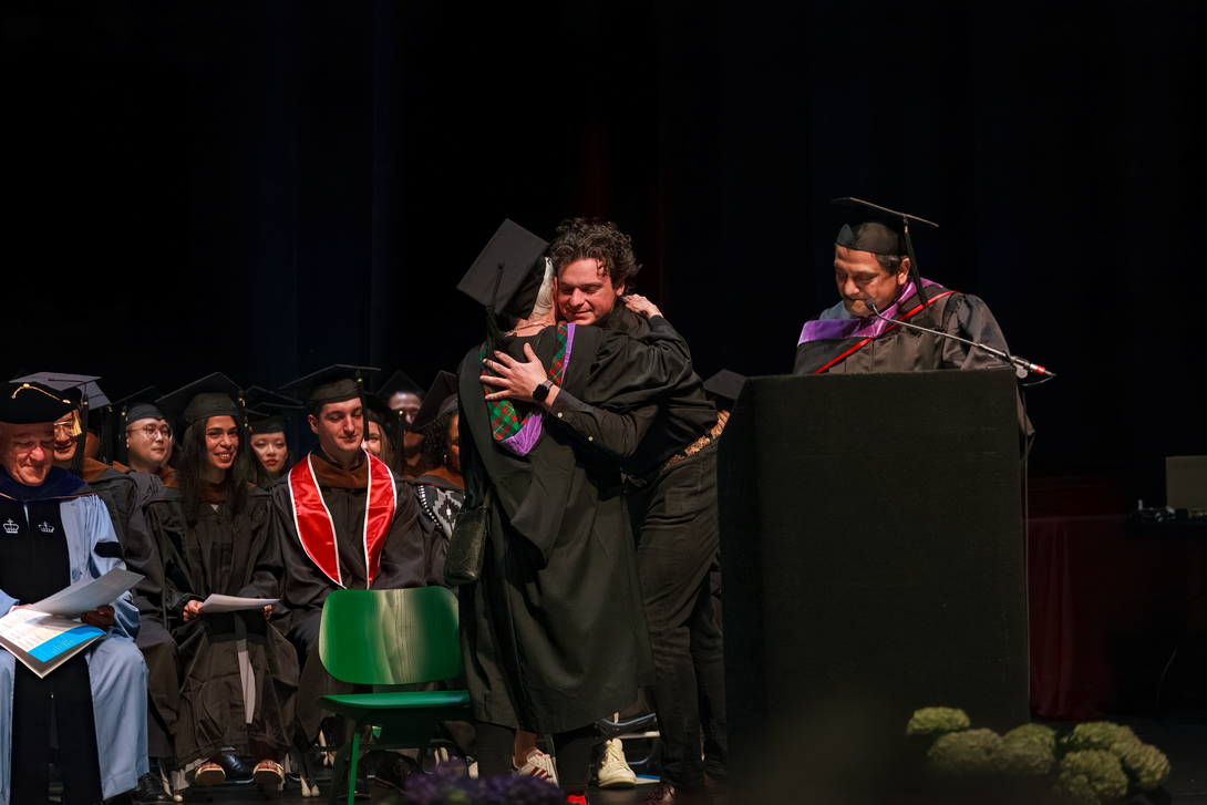 A group of people on stage in graduation regalia. Two people embrace.